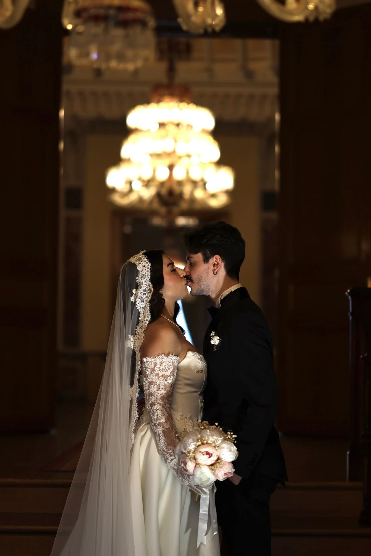 A wedding couple kissing beneath a chandelier, with a lace gown and bouquet